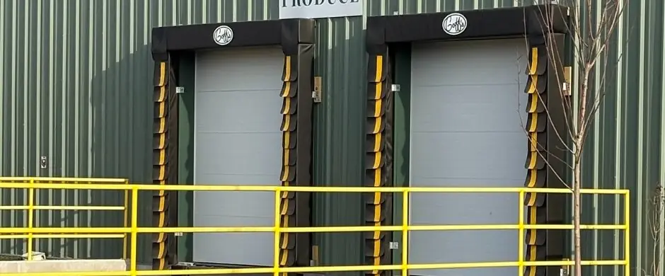 Two loading-dock doors with dock seals and a yellow safety rail on green metal siding at Twin City Produce